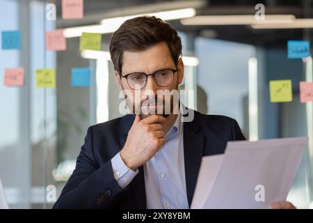 Homme d'affaires mature sérieux profondément concentré sur l'analyse des documents dans le bureau moderne. Port de lunettes et costume de travail. Concept de stratégie commerciale, de planification, de contemplation et de prise de décision. Banque D'Images