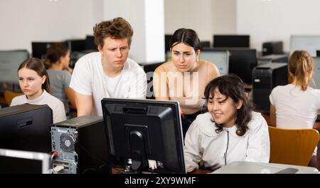 Adolescent assis à table et utilisant l'ordinateur pendant la leçon d'informatique. Professeur et camarades de classe debout et regardant le moniteur Banque D'Images