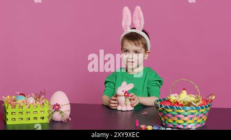 Adorable petit enfant jouant avec un lapin en peluche et un œuf rose à table, s'amusant à décorer des ornements festifs de pâques en studio. Joyeux garçon heureux appréciant les préparations de vacances. Caméra A. Banque D'Images