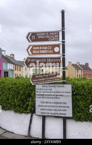 Divers panneaux donnent des directions et des indices dans une rue de maisons colorées et de cieux gris, côte ouest, Portmagee, péninsule d'Iveragh, comté de Kerry, Irelan Banque D'Images