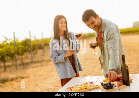 Couple partage le rire et la joie dans un vignoble, savourant de délicieux fromages, fruits et vin au coucher du soleil, jetant une lueur chaude sur leur délicieux p Banque D'Images