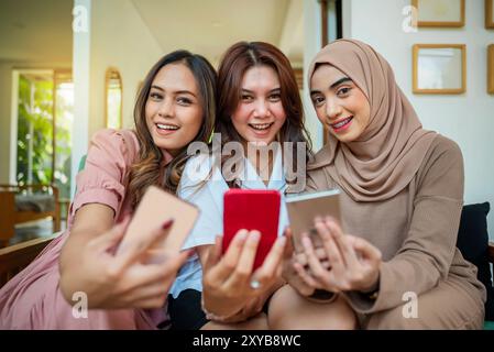 Trois femmes indonésiennes sont assises sur un canapé et passent des appels vidéo avec leur téléphone portable. Ils sourient et semblent s'amuser Banque D'Images