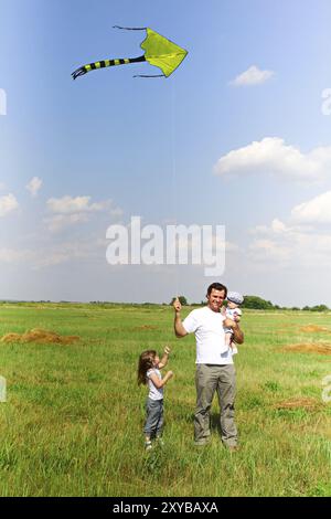 Heureux homme transportant bébé et avec petite fille près de tenir le cerf-volant tout en se tenant sur une pelouse d'été vibrante dans le champ Banque D'Images