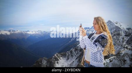 Blonde woman photographing paysage d'hiver montagnes et neige avec cell phone Banque D'Images