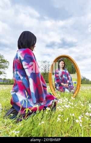 Young colombian woman sits with mirror in flowering pasture Stock Photo