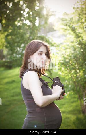 Vue latérale de la charmante jeune femme enceinte dans une robe décontractée prenant des photos avec appareil photo dans le parc vert en été ensoleillé Banque D'Images