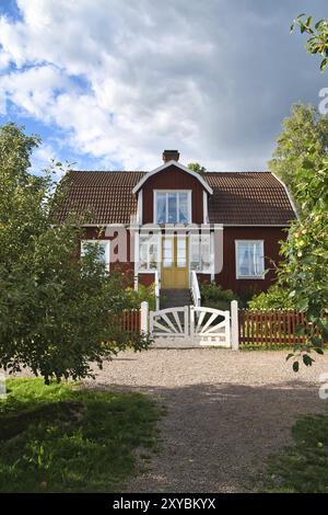 Une maison suédoise rouge et blanche typique dans Smolland. Porte de jardin blanche, clôture brune. pelouse verte dans le jardin. Arbres au premier plan. Ciel bleu avec sm Banque D'Images