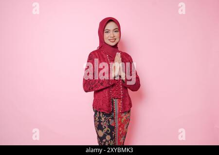 Élégante jeune femme indonésienne portant une robe kebaya moderne et hijab gestuelle saluant ou namaste isolé sur fond rose. Banque D'Images