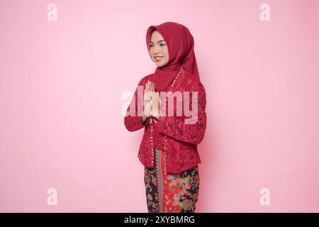 Élégante jeune femme indonésienne portant une robe kebaya moderne et hijab gestuelle saluant ou namaste isolé sur fond rose. Banque D'Images