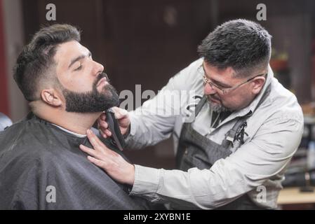 Barbier rasant la barbe d'un bel homme barbu avec un rasoir électrique au salon de coiffure Banque D'Images