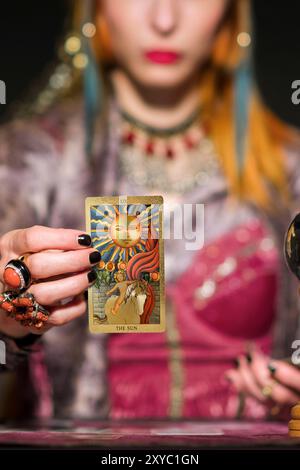 Diseuse de fortune en coiffure de robe lumineuse et anneaux montrant la carte de soleil tout en étant assis à la table pendant la divination dans la chambre sombre Banque D'Images