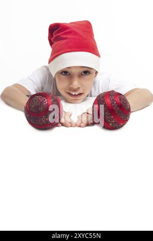 Garçon dans le chapeau rouge de noël tenant des boules décoratives rouges avec une expression heureuse sur son visage avec copyspace blanc pour coller votre annonce ou bannière isolé o Banque D'Images