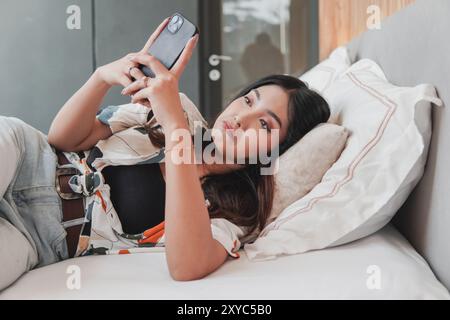 Triste jeune femme asiatique est allongée sur un lit et regarde son smartphone tout en essuyant ses larmes après avoir pleuré. Banque D'Images