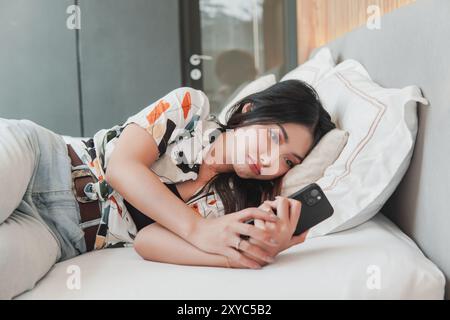 Triste jeune femme asiatique est allongée sur un lit et regarde son smartphone tout en essuyant ses larmes après avoir pleuré. Banque D'Images