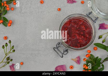 Confiture de framboises dans un pot, décorée de feuilles, brindilles, baies d'orange, pétales de fleurs avec un espace vide à remplir de contenu, vue de dessus. Fond gris Banque D'Images