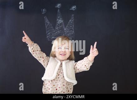 Petite fille mignonne soulevant ses bras et souriant pour l'appareil photo tout en se tenant sous la couronne de craie sur fond noir Banque D'Images