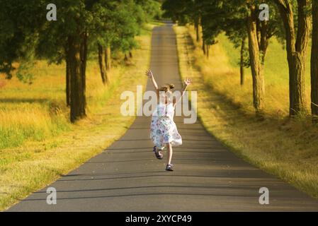 Une petite fille dans une robe d'été court le long d'une rue à la lumière d'une soirée d'été, sautant et gesticulant joyeusement. Une image de la parfaite Happine Banque D'Images