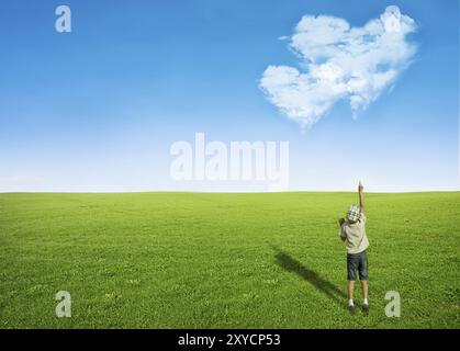 Jeune garçon champ d'herbe verte pointant le ciel bleu avec des nuages en forme de coeur, concept d'amour Banque D'Images