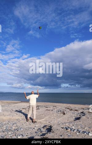 Un homme âgé actif vole un cerf-volant sur une plage en face d'une mer calme et d'un paysage de nuages spectaculaires en été. Un homme fait voler un cerf-volant sur une plage devant un se calme Banque D'Images