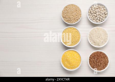 Différents types de céréales et de légumineuses dans des bols sur une table en bois blanc, pose à plat. Espace pour le texte Banque D'Images