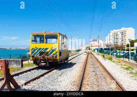 Un train de maintenance jaune sur des voies ferrées près de la côte avec des bâtiments et un parking en arrière-plan sous un ciel bleu vif. Banque D'Images