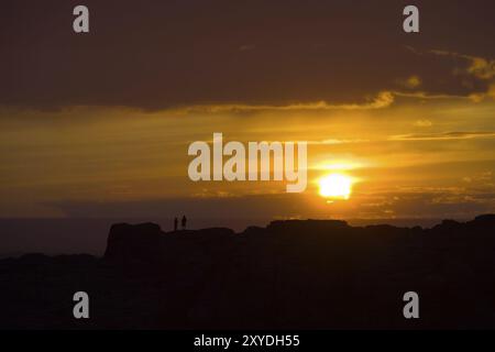 Silhouette of two people standing on distant mountain ridge as setting sun approaches horizon in the Gobi Desert in the countryside of Mongolia Stock Photo