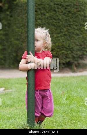 Une petite fille joue sur un poteau de lavage dans le jardin. Elle a l'air malicieuse de derrière le poteau. Petite fille jouant au peek-a-boo dans le peerin du jardin Banque D'Images