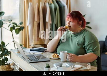 Femme aux cheveux rouges mange des pâtisseries, boit du café, avec ordinateur portable ouvert Banque D'Images