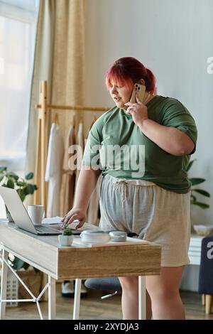 Une femme se tient à son bureau, travaillant sur un ordinateur portable tout en parlant au téléphone. Banque D'Images