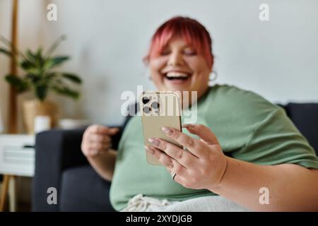 Une femme de grande taille est assise sur un canapé, riant et regardant un smartphone. Banque D'Images