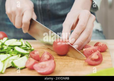 Gros plan les mains féminines délicates coupent un grand couteau avec des tomates sur un quart sur une planche de bois à la maison. Cuisine domestique. Alimentation saine. Le concept de ve Banque D'Images