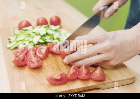 Gros plan les mains féminines délicates coupent un grand couteau avec des tomates sur un quart sur une planche de bois à la maison. Cuisine domestique. Alimentation saine. Le concept de ve Banque D'Images