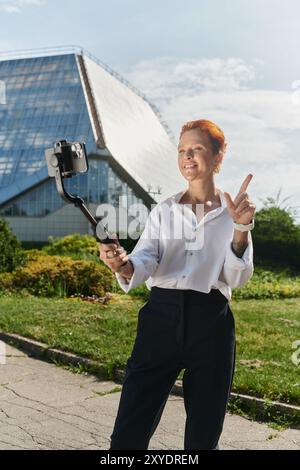 Une femme aux cheveux courts et roux prend un selfie en utilisant un bâton de selfie devant un bâtiment moderne. Banque D'Images