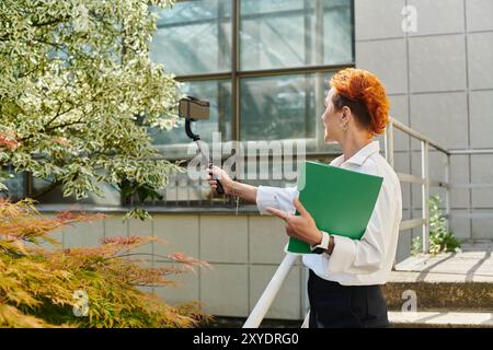 Femme aux cheveux rouges prend un selfie avec un téléphone sur un bâton de selfie tout en tenant un dossier vert Banque D'Images