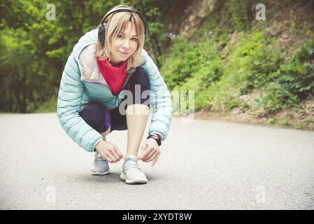 Portrait d'une jeune fille blonde sportive dans des écouteurs sur une course dans la forêt. Une fille assise à lacets noués sur des chaussures de cross-country. Mode de vie actif. Sport Banque D'Images