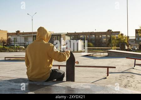 Gros plan d'un jeune hipster vêtu d'un sweat-shirt et d'un Jean assis triste dans un skate Park et tenant une planche à roulettes Banque D'Images