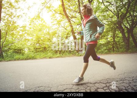 Jeune femme blonde de forme physique dans un casque courant le matin sentier de forêt caucasienne à la lumière du soleil Banque D'Images