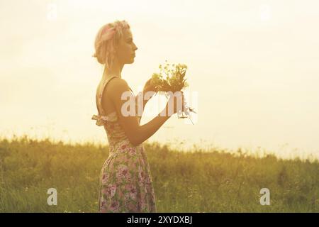 Jeune femme vêtue avec bouquet de fleurs entre les mains au coucher du soleil dans le champ. Image teintée et chaude Banque D'Images