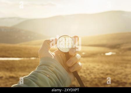 La main d'un homme tient une boussole à main sur fond de montagnes et de collines au coucher du soleil. Le concept de voyage et de navigation dans les espaces ouverts Banque D'Images