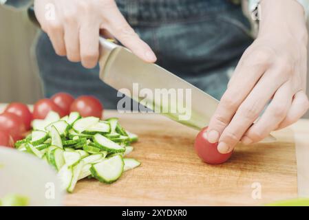 Gros plan les mains féminines délicates coupent un grand couteau avec des tomates sur un quart sur une planche de bois à la maison. Cuisine domestique. Alimentation saine. Le concept de ve Banque D'Images