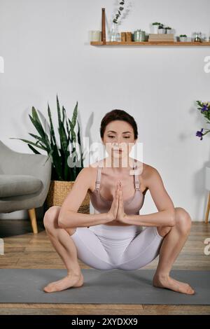 Une femme dans un crop top et un short médite en position accroupie sur un tapis de yoga dans son appartement moderne. Banque D'Images