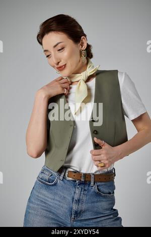 Une femme asiatique dans un gilet vert, un haut blanc, une écharpe jaune clair et une ceinture brune pose sur un fond gris avec des cheveux dans un chignon bas Banque D'Images