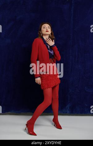 Une femme en costume rouge, chemise en soie et bas pose contre un mur bleu. Banque D'Images