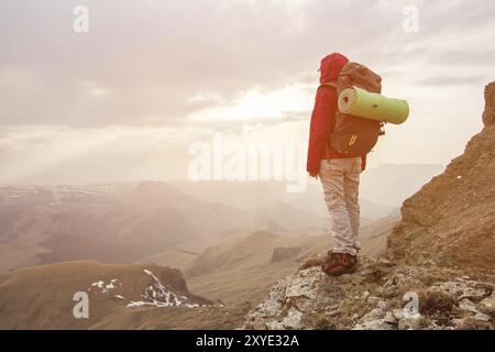 Fille voyageuse dans une veste rouge avec un sac à dos et dans une casquette regarde assis sur un rocher haut dans les montagnes du caucase sur un soleil couchant sur le rocher Banque D'Images