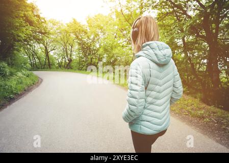 Portrait d'une fille blonde athlétique attrayante de dos à la lumière courir vers le bas veste, portant des écouteurs bluetooth avec de la musique ou des sons de la nature quand o Banque D'Images