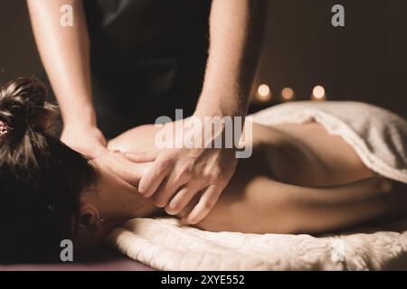 Les mains des hommes font un massage thérapeutique du cou pour une fille allongée sur un canapé de massage dans un spa de massage avec un éclairage sombre. Gros plan. Dark Key Banque D'Images