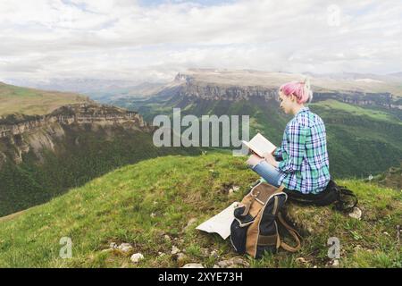 Une fille voyageuse est assise dans les montagnes sur l'herbe et lit un livre sur le fond de montagnes épiques. Le concept de lecture pendant le repos et vacati Banque D'Images