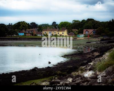 Des maisons colorées bordent le littoral, reflétant dans des eaux calmes. La plage rocheuse et les vagues douces créent une atmosphère tranquille sous un ciel nuageux. Banque D'Images