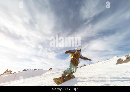 Un snowboarder dans un masque de ski et un sac à dos roule sur une pente enneigée laissant derrière lui une poudre de neige contre le ciel bleu et le soleil couchant. Banque D'Images