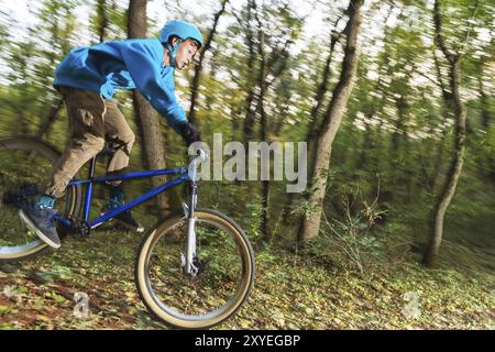 Un jeune cavalier dans un casque et un sweat-shirt bleu vole sur un vélo après avoir sauté d'un haut kicker sur une piste cyclable forestière. Prise de vue avec exposition prolongée Banque D'Images